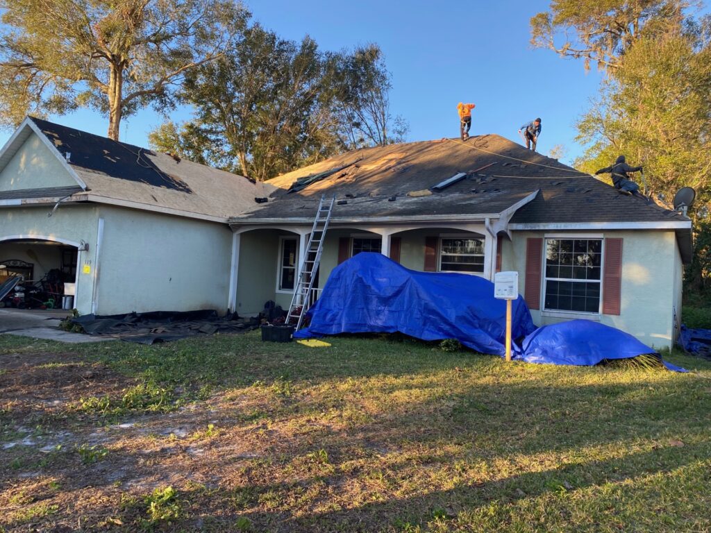 Roofers in safety harnesses tearing off old shingles from a stucco home in DeLand, FL, with blue tarps protecting the landscaping, before installing Atlas Pinnacle Pristine shingles in Majestic Shake.