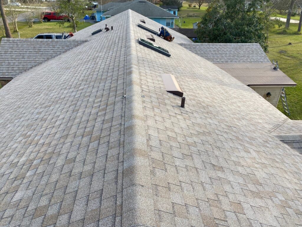Drone view of a hip roof in St. Augustine, FL with tan Atlas Pinnacle Sun architectural shingles, skylights, roof vents, and a roofer working near the ridge.