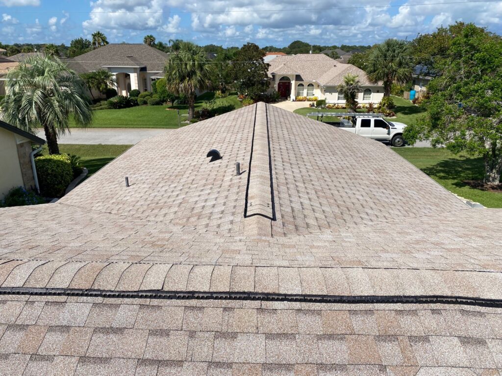 New Atlas Pinnacle Pristine shingles in Tan on a hip roof in Palm Coast, FL, with ridge vent being installed and palm trees and neighborhood homes in the background.